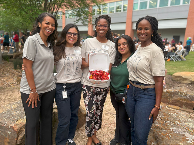 UTHealth Houston Student Affairs Team smiling outdoors during a campus crawfish boil; one holds a takeout container filled with crawfish.
